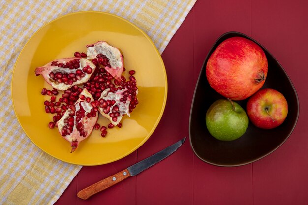 Draufsicht von Granatapfelstücken und -beeren im Teller auf kariertem Stoff mit Granatapfelapfel und -messer auf Bordooberfläche