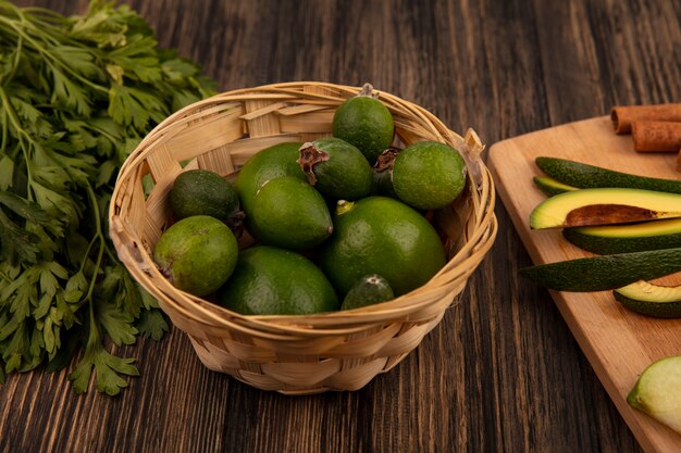 Draufsicht von grünen reifen Feijoas auf einem Eimer mit gehackten Scheiben von Avocados auf einem hölzernen Küchenbrett mit Petersilie lokalisiert auf einem hölzernen Hintergrund