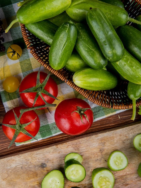 Draufsicht von Gurken im Korb mit Tomaten auf Stoff und Gurkenscheiben auf Holz