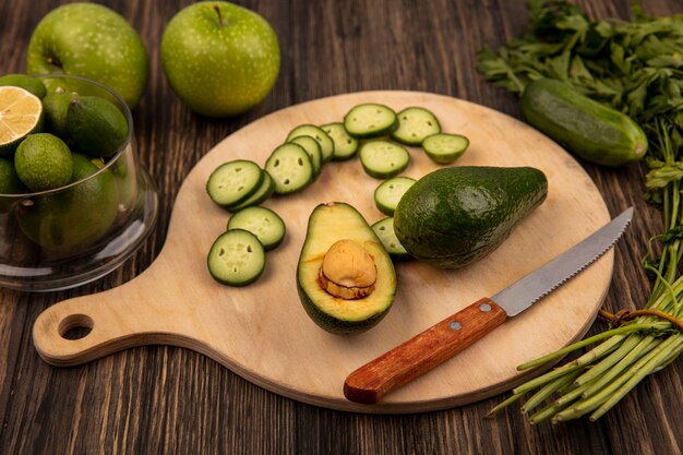 Draufsicht von Gurkenscheiben auf einem hölzernen Küchenbrett mit Avocado mit Messer mit Feijoas auf einer Glasschale mit grünen Äpfeln und Petersilie lokalisiert auf einer Holzoberfläche