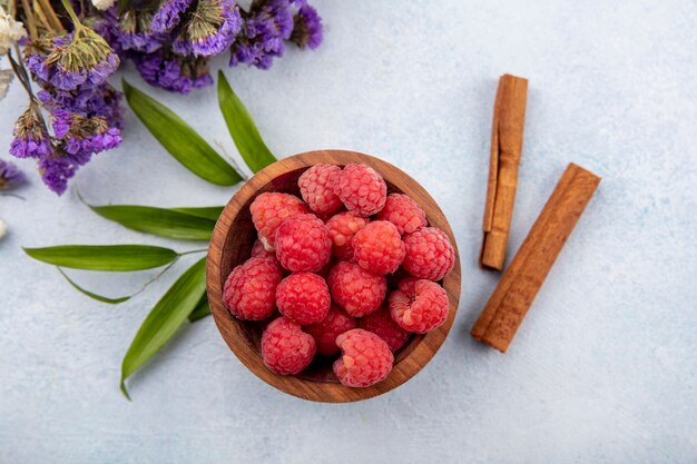 Draufsicht von Himbeeren in Schüssel und Zimt mit Blumen und Blättern auf weißer Oberfläche