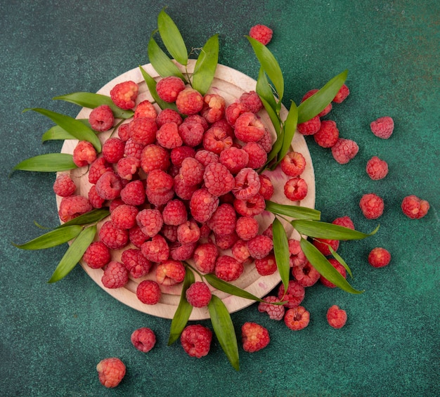 Draufsicht von Himbeeren mit Blättern auf Schneidebrett auf grüner Oberfläche