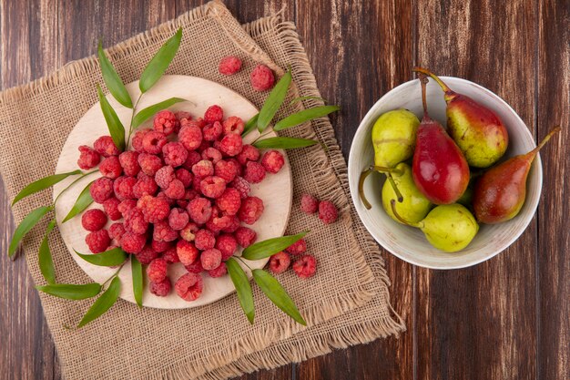 Draufsicht von Himbeeren mit Blättern auf Schneidebrett auf Sack und Pfirsichschale auf Holzoberfläche