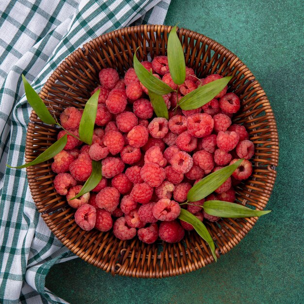 Draufsicht von Himbeeren mit Blättern im Korb auf kariertem Stoff und auf grüner Oberfläche