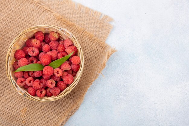 Draufsicht von Himbeeren mit Blättern im Korb auf Sackleinen und auf weißer Oberfläche