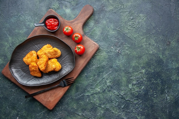 Draufsicht von Hühnernuggets auf einem schwarzen Teller und einer Gabel auf Holzbrett-Tomatenketchup auf der rechten Seite auf dunkler Oberfläche