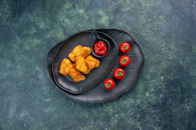 Draufsicht von Hühnernuggets und Ketchup-Tomaten in schwarzen Tellern auf dunkler Oberfläche