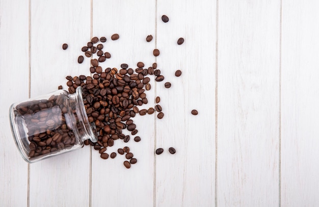 Draufsicht von Kaffeebohnen, die aus einem Glas auf einem weißen hölzernen Hintergrund mit Kopienraum fallen