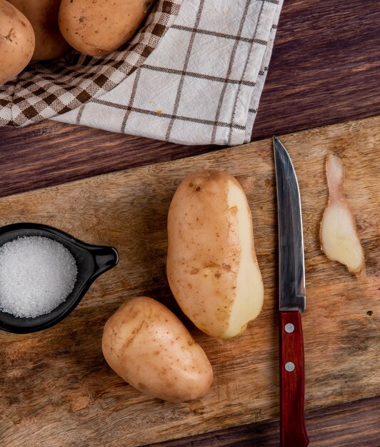 Draufsicht von Kartoffeln mit Muschelmesser und Salz auf Schneidebrett mit anderen im Korb auf Stoff auf Holzoberfläche