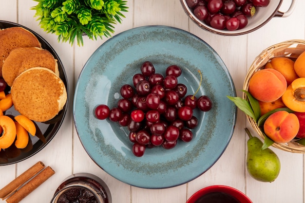 Draufsicht von Kirschen in Teller und Pfannkuchen Aprikosenscheiben in Teller mit Korb von Aprikose und Marmelade Zimtbirne auf Holzhintergrund