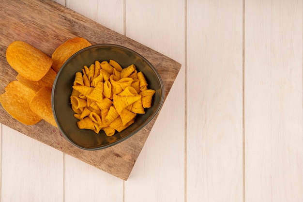 Draufsicht von leckeren knusprigen Kegelform-Maissnacks auf einer Schüssel auf einem hölzernen Küchenbrett auf einem beigen Holztisch mit Kopierraum