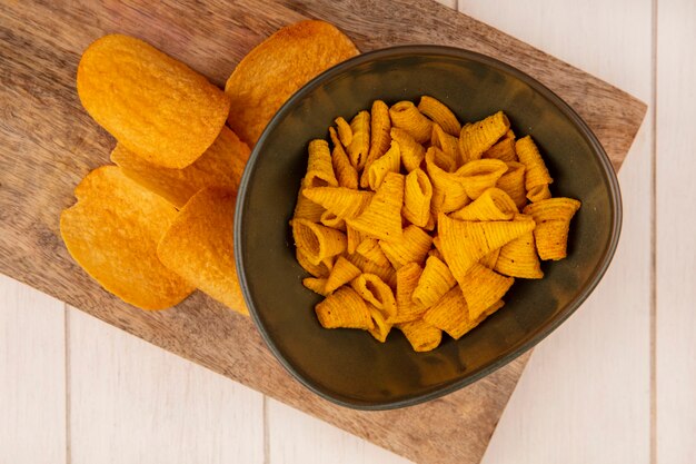 Draufsicht von leckeren knusprigen Kegelform-Maissnacks auf einer Schüssel auf einem hölzernen Küchenbrett auf einem beigen Holztisch