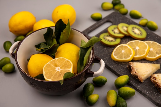 Draufsicht von leckeren Scheiben von Früchten wie Kiwi-Ananas-Zitrone auf einem schwarzen Küchenbrett mit Zitronen auf einer Schüssel mit Kinkans und Zitronen isoliert auf einer grauen Wand