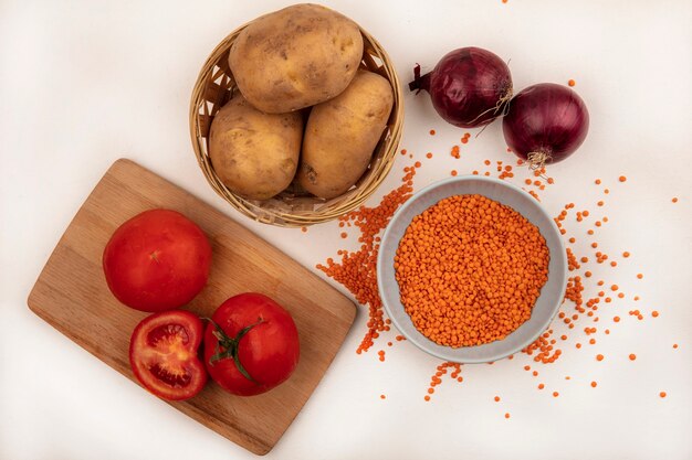 Draufsicht von leuchtend orangefarbenen Linsen auf einer Schüssel mit Kartoffeln auf einem Eimer mit Tomaten auf einem hölzernen Küchenbrett mit roten Zwiebeln lokalisiert auf einer weißen Wand