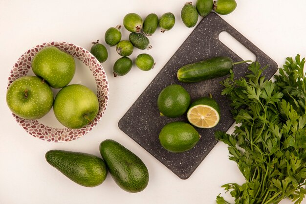 Draufsicht von Limetten auf einem Küchenbrett mit Äpfeln auf einer Schüssel mit Gurkenfeijoas und Avocados lokalisiert auf einer weißen Wand