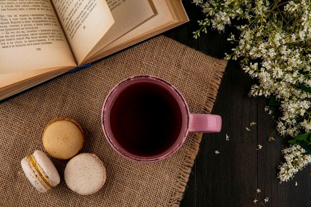 Draufsicht von Macarons mit einer Tasse Tee auf einer beigen Serviette mit einem offenen Buch und Blumen