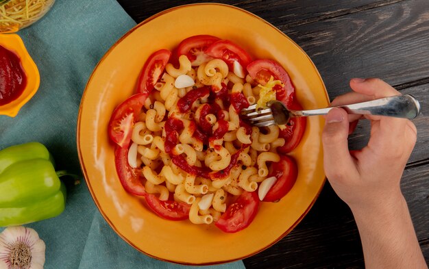 Draufsicht von Makkaroni-Nudeln in Platte mit Pfeffer-Ketchup-Knoblauch und Handhaltegabel auf blauem Stoff und Holzoberfläche