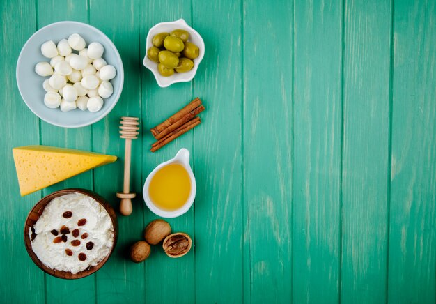 Draufsicht von Mozzarella-Käse in einer Schüssel Hüttenkäse und einem Stück holländischem Käse mit Walnusshonig-Zimtstangen und eingelegten Oliven auf grünem Holz mit Kopierraum