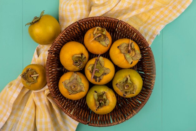 Draufsicht von orange unreifen Kakifrüchten auf einem Eimer auf einem gelben karierten Tuch auf einem blauen Holztisch