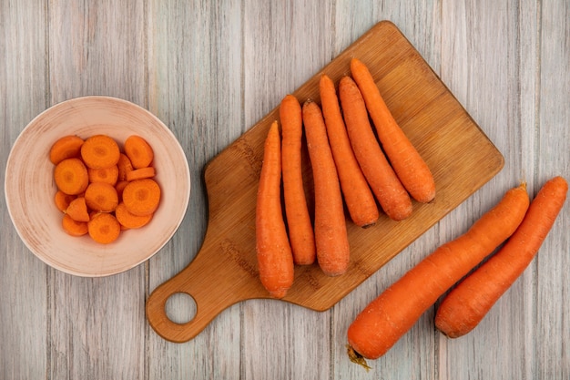 Draufsicht von Orangenwurzelgemüse-Karotten auf einem hölzernen Küchenbrett mit gehackten Karotten auf einer Schüssel auf einer grauen Holzoberfläche