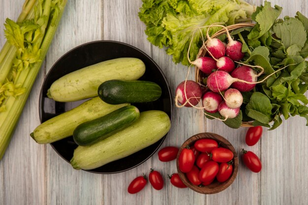 Draufsicht von organischen Gurken und Zucchini auf einem Teller mit Tomaten auf einer Holzschale mit Radieschen auf einem Eimer mit Salattomaten und Sellerie lokalisiert auf einer grauen Holzoberfläche