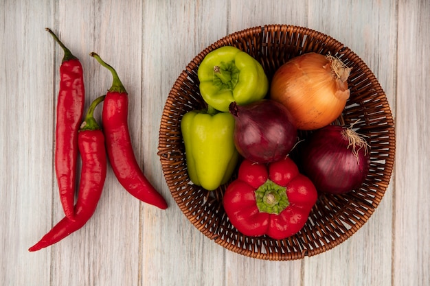 Draufsicht von Paprika auf einem Eimer mit Zwiebeln mit Chilischoten lokalisiert auf einem grauen hölzernen Hintergrund