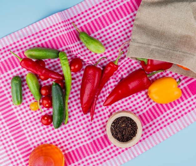 Draufsicht von Paprika, die aus dem Sack mit Gurkentomaten und schwarzen Pfeffersamen mit geschmolzenem Öl auf kariertem Stoff und blauer Oberfläche verschüttet werden