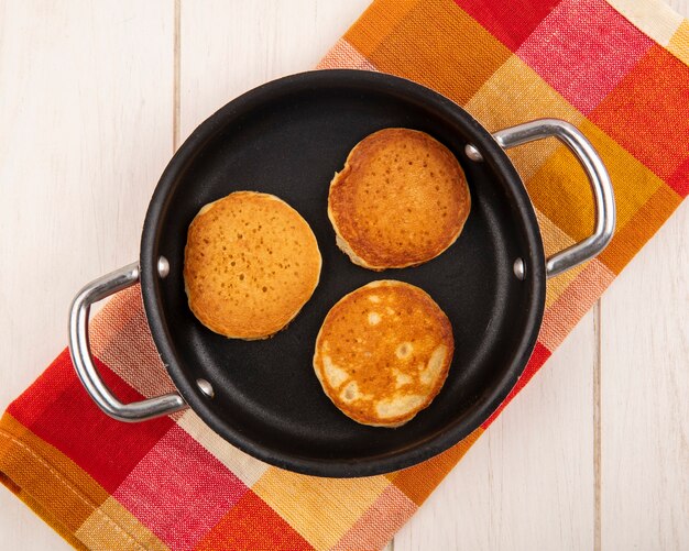 Draufsicht von Pfannkuchen in der Pfanne auf kariertem Stoff und hölzernem Hintergrund