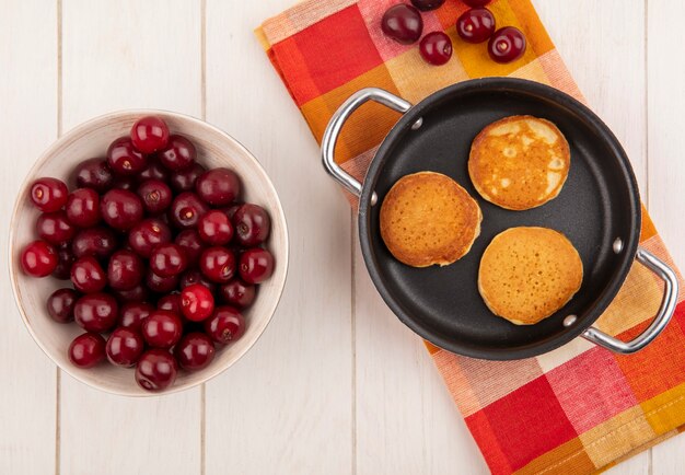 Draufsicht von Pfannkuchen in der Pfanne und in den Kirschen auf kariertem Stoff und in der Schüssel auf hölzernem Hintergrund