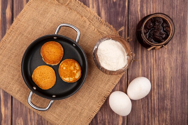 Draufsicht von Pfannkuchen in Pfanne und Mehl in Schüssel auf Sackleinen mit Eiern und Erdbeermarmelade auf hölzernem Hintergrund