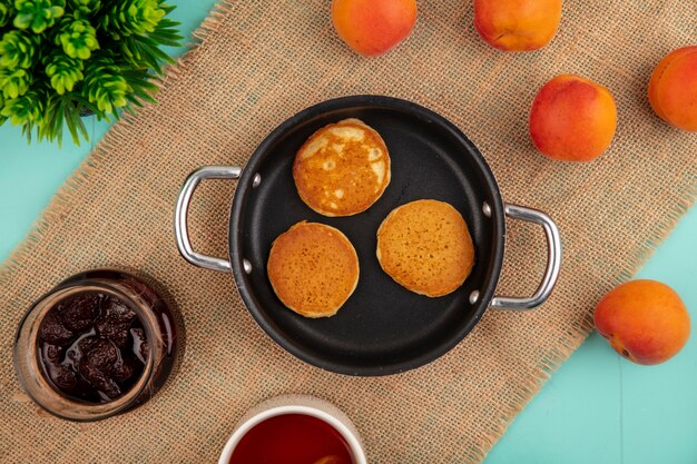 Draufsicht von Pfannkuchen in Pfannen- und Erdbeermarmeladen-Tee-Aprikosen auf Sackleinen auf blauem Hintergrund