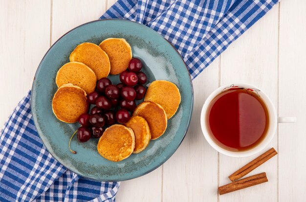Draufsicht von Pfannkuchen mit Kirschen in Platte auf kariertem Stoff und Tasse Tee mit Zimt auf hölzernem Hintergrund