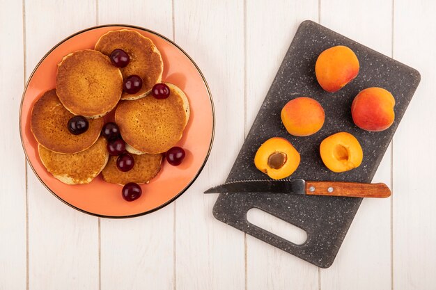 Draufsicht von pfannkuchen mit kirschen in platte und aprikosen mit messer auf schneidebrett auf hölzernem hintergrund