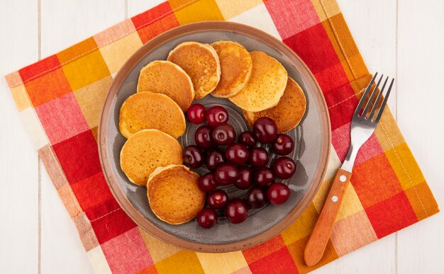 Draufsicht von Pfannkuchen mit Kirschen in Platte und Gabel auf kariertem Stoff und hölzernem Hintergrund