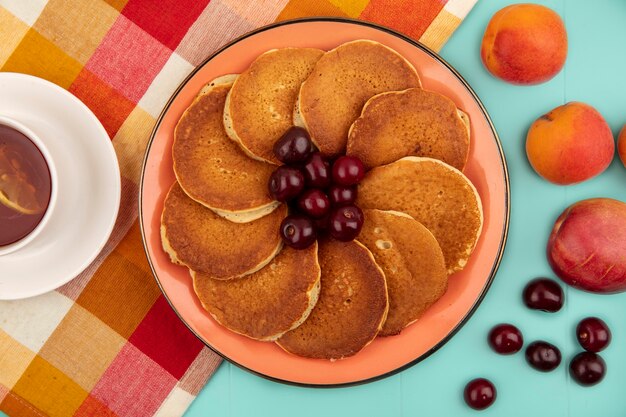 Draufsicht von Pfannkuchen mit Kirschen in Teller und Tasse Tee auf kariertem Stoff mit Aprikosen auf blauem Hintergrund