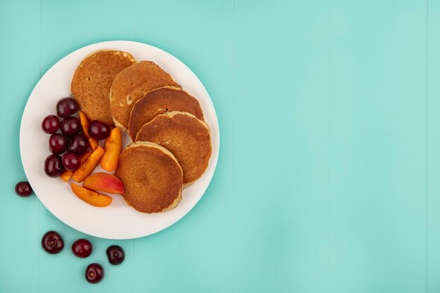 Draufsicht von Pfannkuchen mit Kirschen und Aprikosenscheiben in Platte auf blauem Hintergrund mit Kopienraum