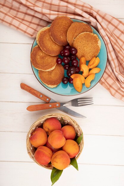 Draufsicht von Pfannkuchen mit Kirschen und Aprikosenstücken in Platte auf kariertem Stoff und Korb von Aprikosen mit Gabel und Messer auf hölzernem Hintergrund