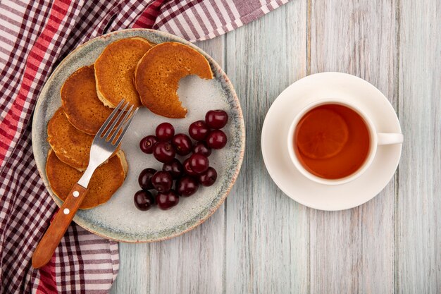 Draufsicht von Pfannkuchen mit Kirschen und Gabel in Platte auf kariertem Stoff und Tasse Tee auf hölzernem Hintergrund