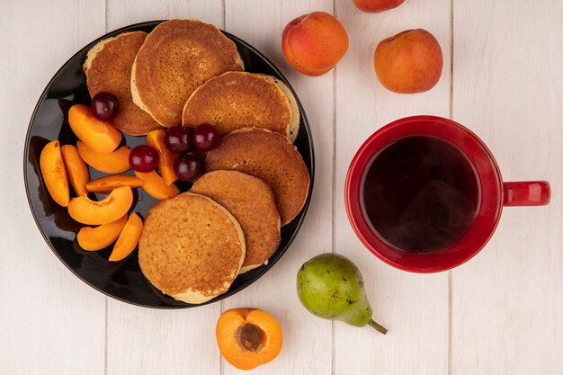 Draufsicht von Pfannkuchen mit Kirschen und geschnittener Aprikose in Teller und Tasse Kaffee mit Birnen und Aprikosen auf hölzernem Hintergrund