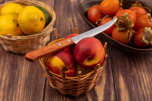 Draufsicht von Pfirsichen auf einem Eimer mit Messer mit Mandarinen mit Kakis auf einer Schüssel auf einer Holzoberfläche