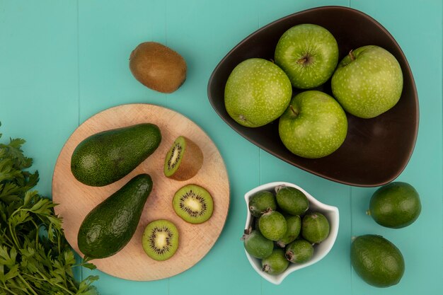 Draufsicht von reifen Avocados mit Kiwischeiben auf einem hölzernen Küchenbrett mit Feijoas auf einer Schüssel mit Äpfeln auf einer Schüssel mit Limetten lokalisiert auf einer blauen Wand