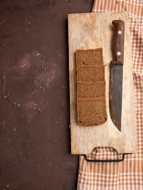 Draufsicht von Roggenbrotscheiben und Messer auf Schneidebrett auf Stoff auf kastanienbraunem Hintergrund mit Kopienraum