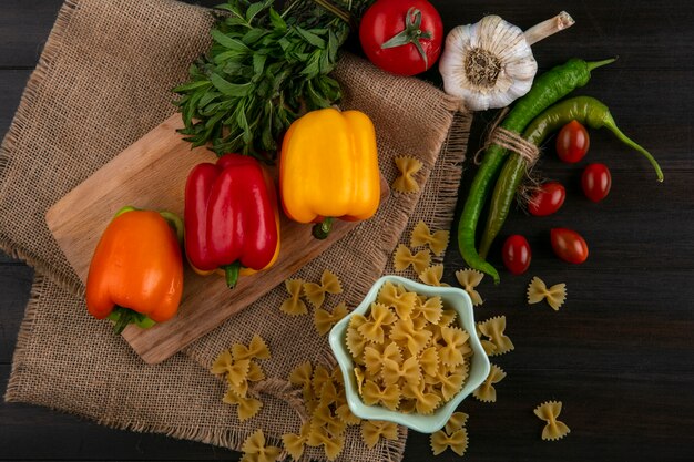 Draufsicht von rohen Nudeln mit farbigen Paprika auf einem Schneidebrett mit Knoblauch-Tomaten-Chilischoten und Minze auf einer beigen Serviette