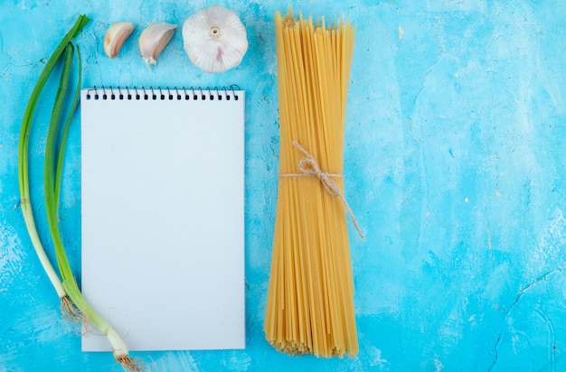 Draufsicht von rohen spaghetti gebunden mit seil knoblauch skizzenbuch und frühlingszwiebeln mit kopienraum auf blauem hintergrund
