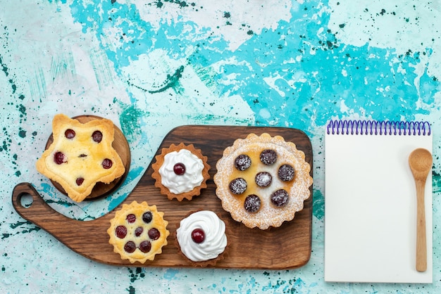 Draufsicht von Sahnekuchen mit Früchten zusammen mit Notizblock auf Graulichtschreibtisch, Kuchenkeks süßer Zucker