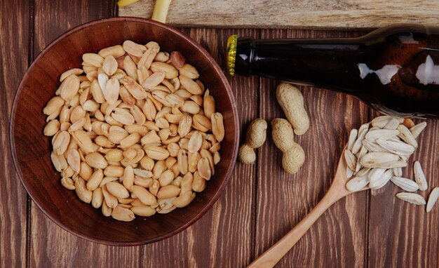 Draufsicht von salzigen Snack-Erdnüssen in einer Holzschale mit Sonnenblumenkernen in einem Holzlöffel und einer Flasche Bier auf rustikalem