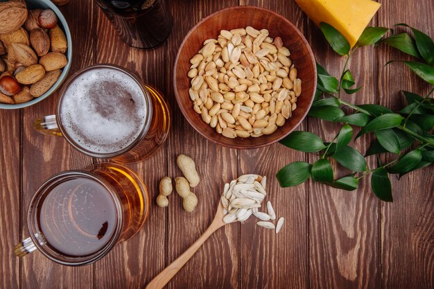 Draufsicht von salzigen Snack-Erdnüssen in einer Schüssel und Sonnenblumenkernen in einem Holzlöffel mit zwei Bechern Bier auf rustikalem Holz