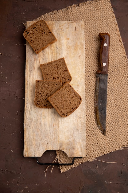 Draufsicht von Schwarzbrotscheiben auf Schneidebrett mit Messer auf Sackleinen auf kastanienbraunem Hintergrund