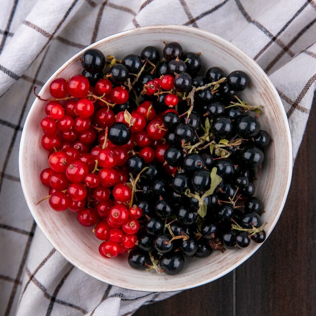 Draufsicht von schwarzen und roten Johannisbeeren in einer Schüssel auf einem karierten Handtuch auf einer Holzoberfläche