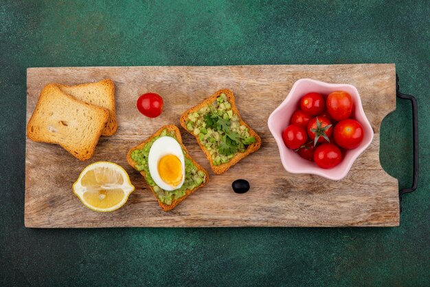 Draufsicht von Tomaten auf einer rosa Schüssel auf einem hölzernen Küchenbrett mit gerösteten Brotscheiben mit Avocadopulpe und Ei auf grüner Oberfläche